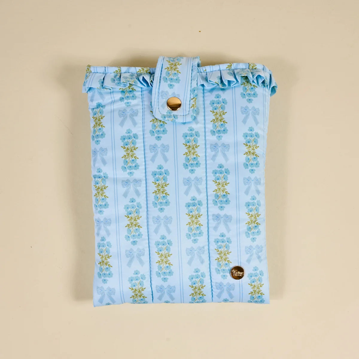 A light blue fabric pouch with a vertical striped pattern of blue bows and yellow flowers, featuring a ruffled top edge and a snap-button closure, laid flat on a beige background.