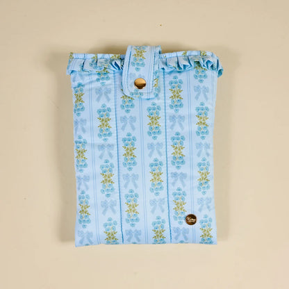 A light blue fabric pouch with a vertical striped pattern of blue bows and yellow flowers, featuring a ruffled top edge and a snap-button closure, laid flat on a beige background.