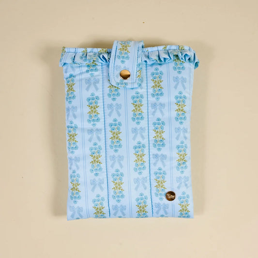 A light blue fabric pouch with a vertical striped pattern of blue bows and yellow flowers, featuring a ruffled top edge and a snap-button closure, laid flat on a beige background.