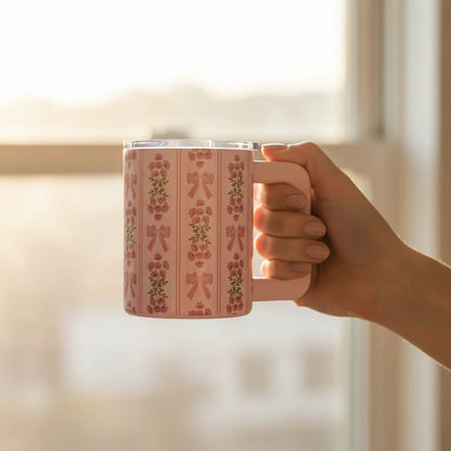 A hand holding a pink ceramic mug with a vertical pattern of pink bows, cherries, and small green floral motifs, set against a softly blurred warm-toned window background.