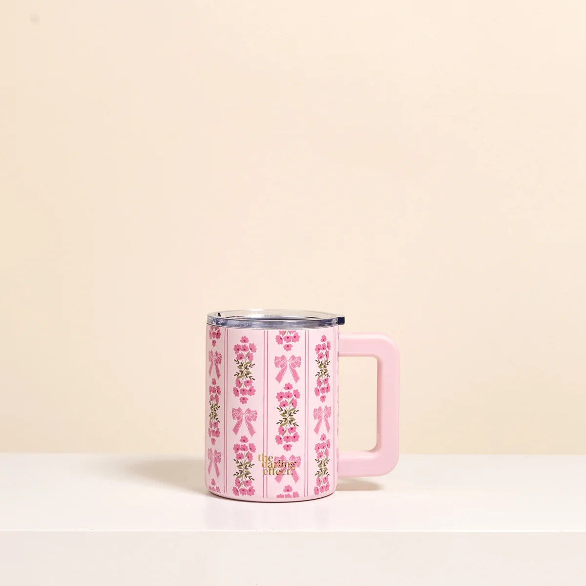 A pink handled mug with a floral and bow pattern, featuring a clear lid, set against a minimalist beige background.