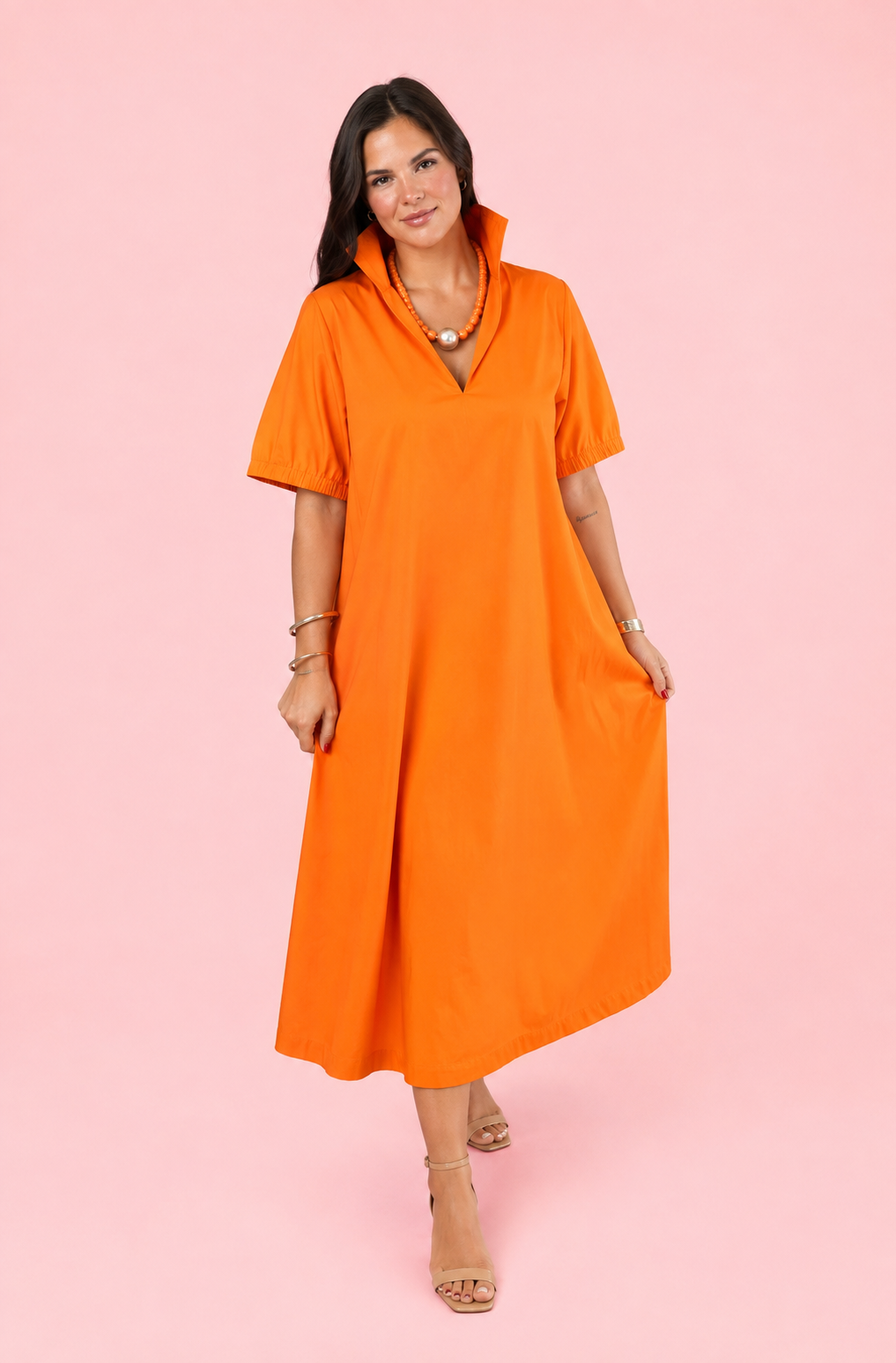 A woman modeling a bright orange short-sleeve midi dress with a high collar and split neckline, standing against a solid pink studio background.