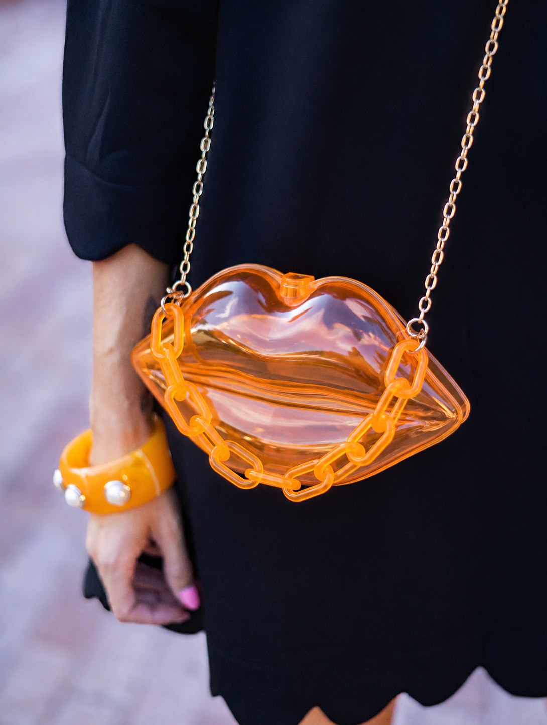 A close-up of a person wearing a black dress and orange bracelet, carrying a transparent orange lips-shaped shoulder bag with a gold chain strap.