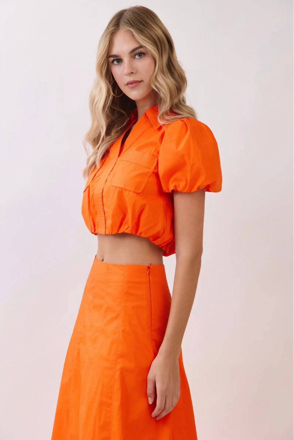 A woman modeling a bright orange two-piece outfit consisting of a cropped puff-sleeve top and a matching high-waisted A-line skirt, set against a plain light beige studio background.