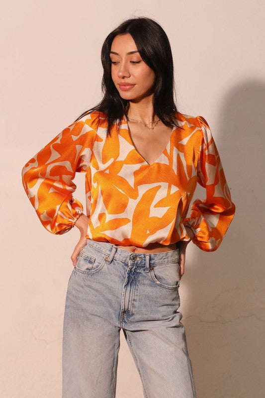 A woman wearing a vibrant orange and white abstract patterned blouse with voluminous sleeves, paired with light-wash high-waisted jeans, standing against a plain beige wall.