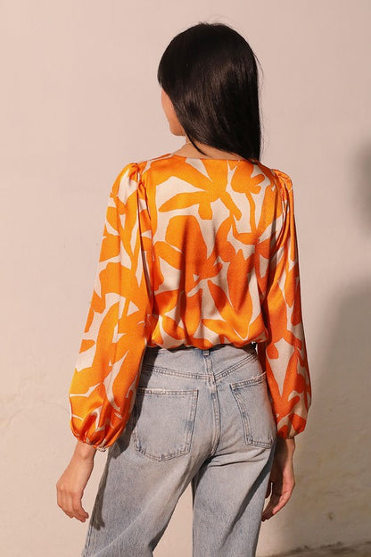 A woman shown from the back wearing a long-sleeve blouse with an orange and cream abstract floral pattern, paired with light-wash jeans, against a plain beige wall.