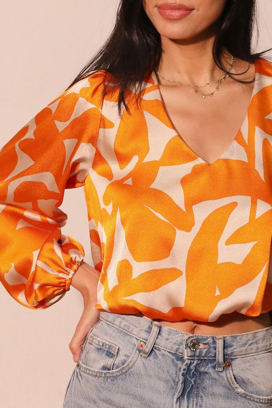 A woman wearing a vibrant orange and cream abstract patterned blouse with long balloon sleeves and a V-neckline, paired with light-wash blue jeans.