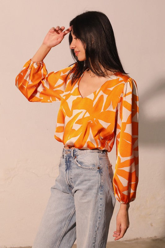 A woman wearing a vibrant orange and white abstract patterned blouse with voluminous long sleeves, paired with light-wash blue jeans, posing against a neutral beige wall.