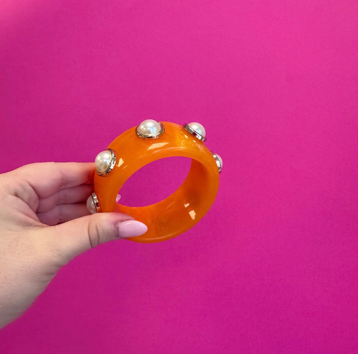 An orange translucent bangle bracelet with silver-toned metal studs and white pearl-like accents, held by a hand against a solid bright pink background.