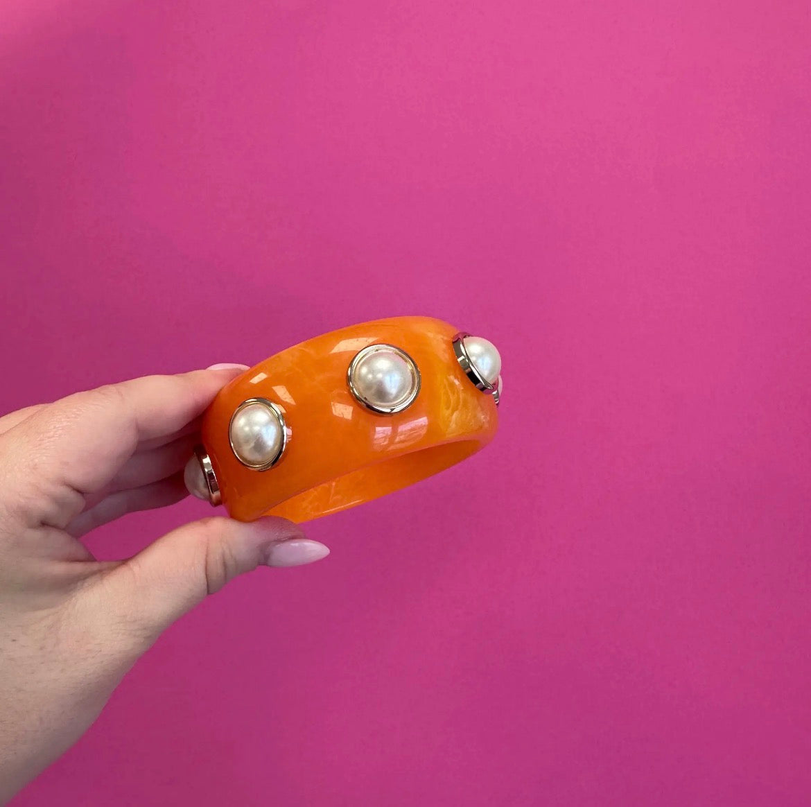 An orange resin cuff bracelet with three faux pearl studs in gold bezels, held by a hand against a solid pink background.