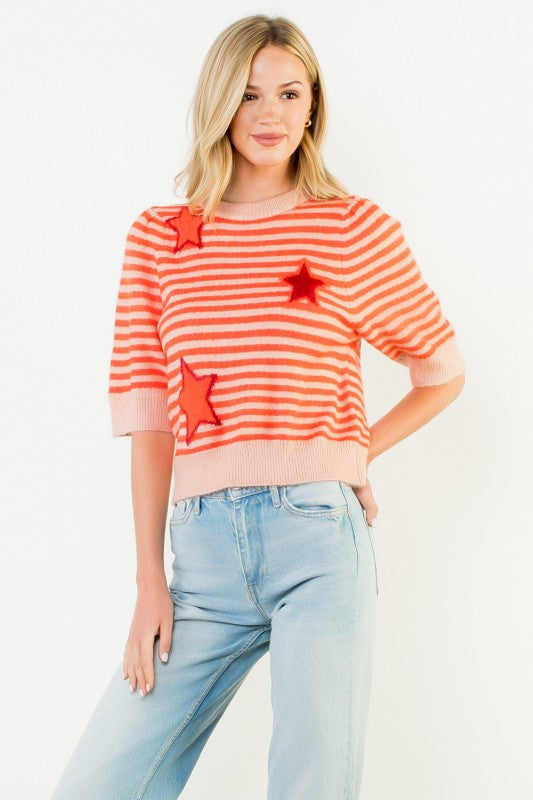 A woman modeling a short-sleeve knit sweater with horizontal orange and white stripes, accented with three red star appliqués, paired with light blue jeans against a plain white background.