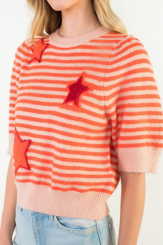 A woman wearing a fuzzy knit sweater with horizontal orange and cream stripes, decorated with three dimensional red sequined stars, paired with light blue jeans.