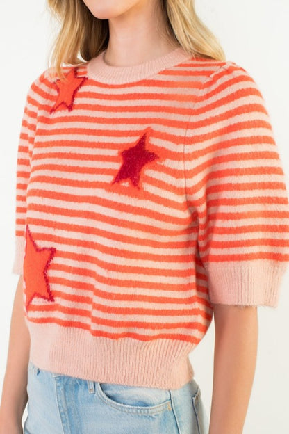 A woman wearing a fuzzy knit sweater with horizontal orange and cream stripes, decorated with three dimensional red sequined stars, paired with light blue jeans.