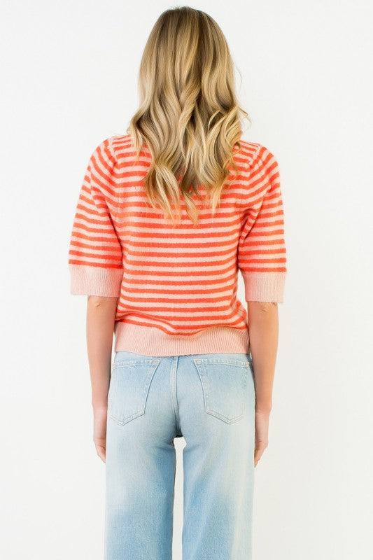 A woman viewed from behind wearing a short-sleeve knit top with horizontal coral-orange and white stripes, paired with light blue jeans, against a plain white background.