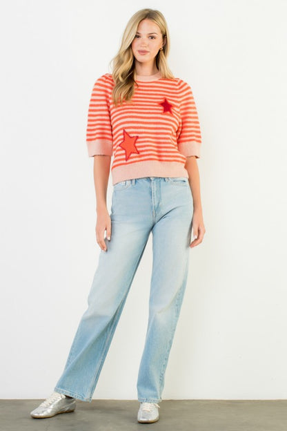 A woman modeling an orange pink and orange striped knit sweater with red star patches, paired with light blue jeans and silver sneakers, against a plain white studio background.