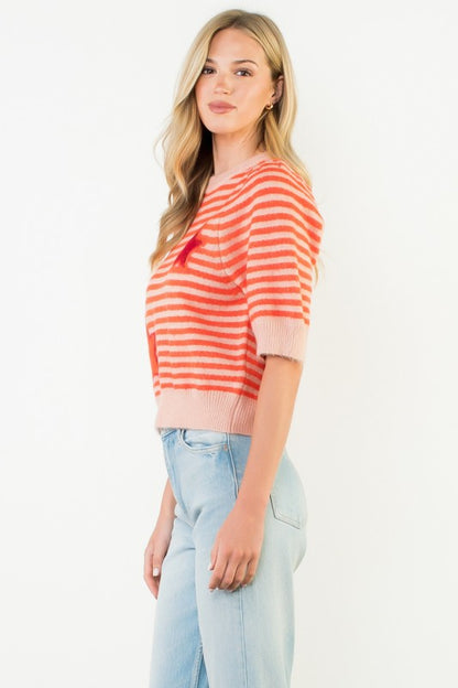 A woman modeling a short-sleeve knit top with horizontal coral and pink stripes, paired with light blue jeans, against a plain white background.