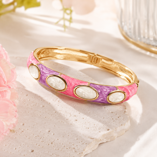 A gold-plated bangle bracelet with a pink and purple marbled enamel body and four white oval cabochon stones, displayed on a textured stone slab.