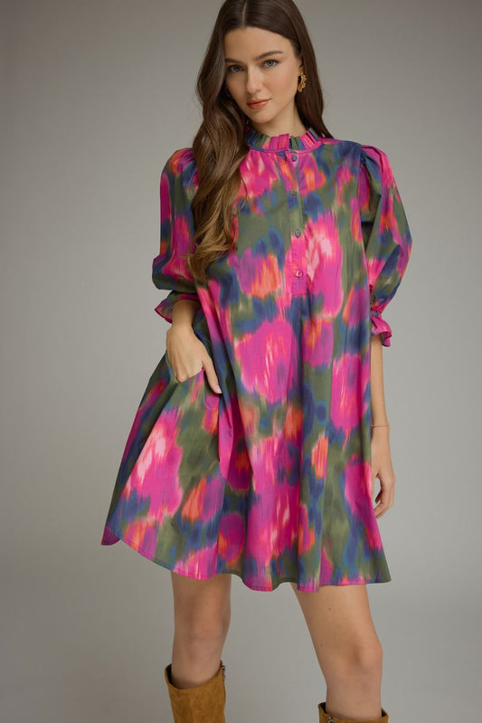 A woman modeling a short puff-sleeve dress with a vibrant pink, purple, and green abstract tie-dye pattern, paired with brown boots, against a solid gray studio background.
