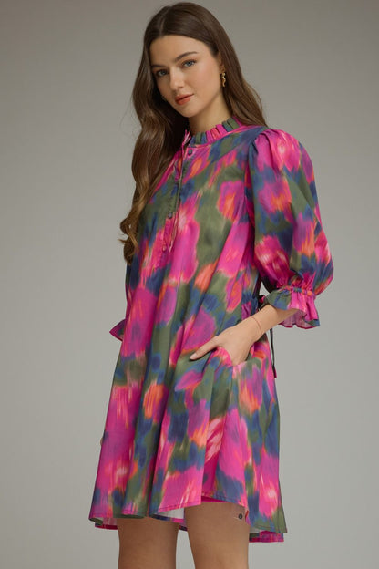 A woman modeling a short puff-sleeve dress with a vibrant pink, purple, and green abstract tie-dye pattern against a solid gray studio background.