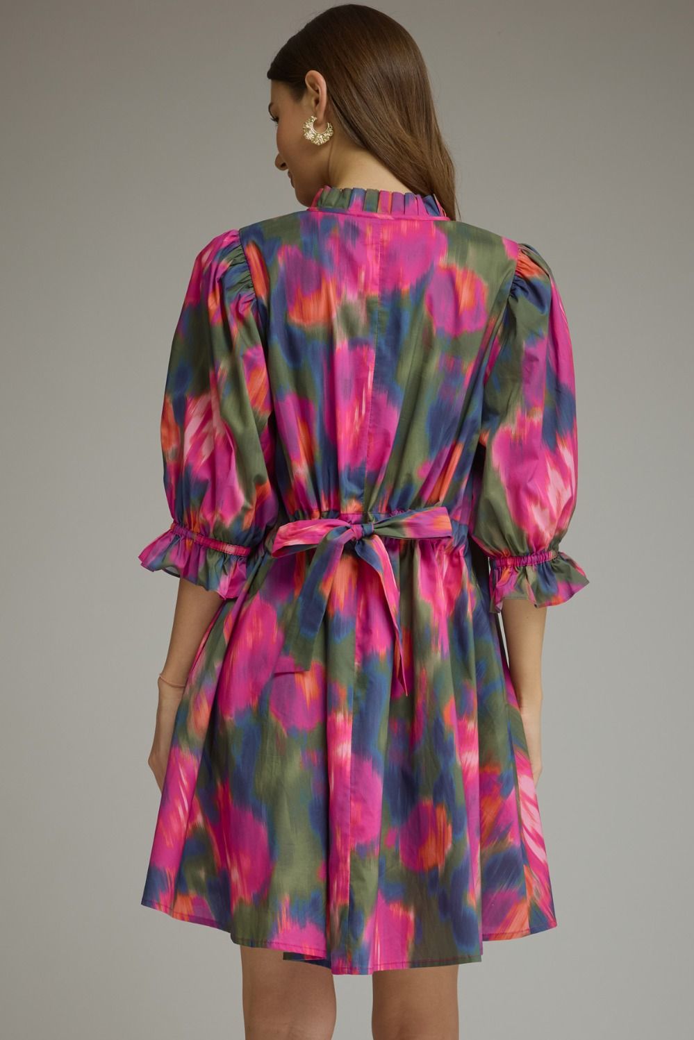 A woman shown from the back wearing a short puff-sleeve dress with a tie waist, featuring a vibrant abstract floral pattern in pink, green, and blue on a light gray background.