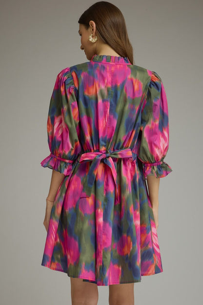 A woman shown from the back wearing a short puff-sleeve dress with a tie waist, featuring a vibrant abstract floral pattern in pink, green, and blue on a light gray background.