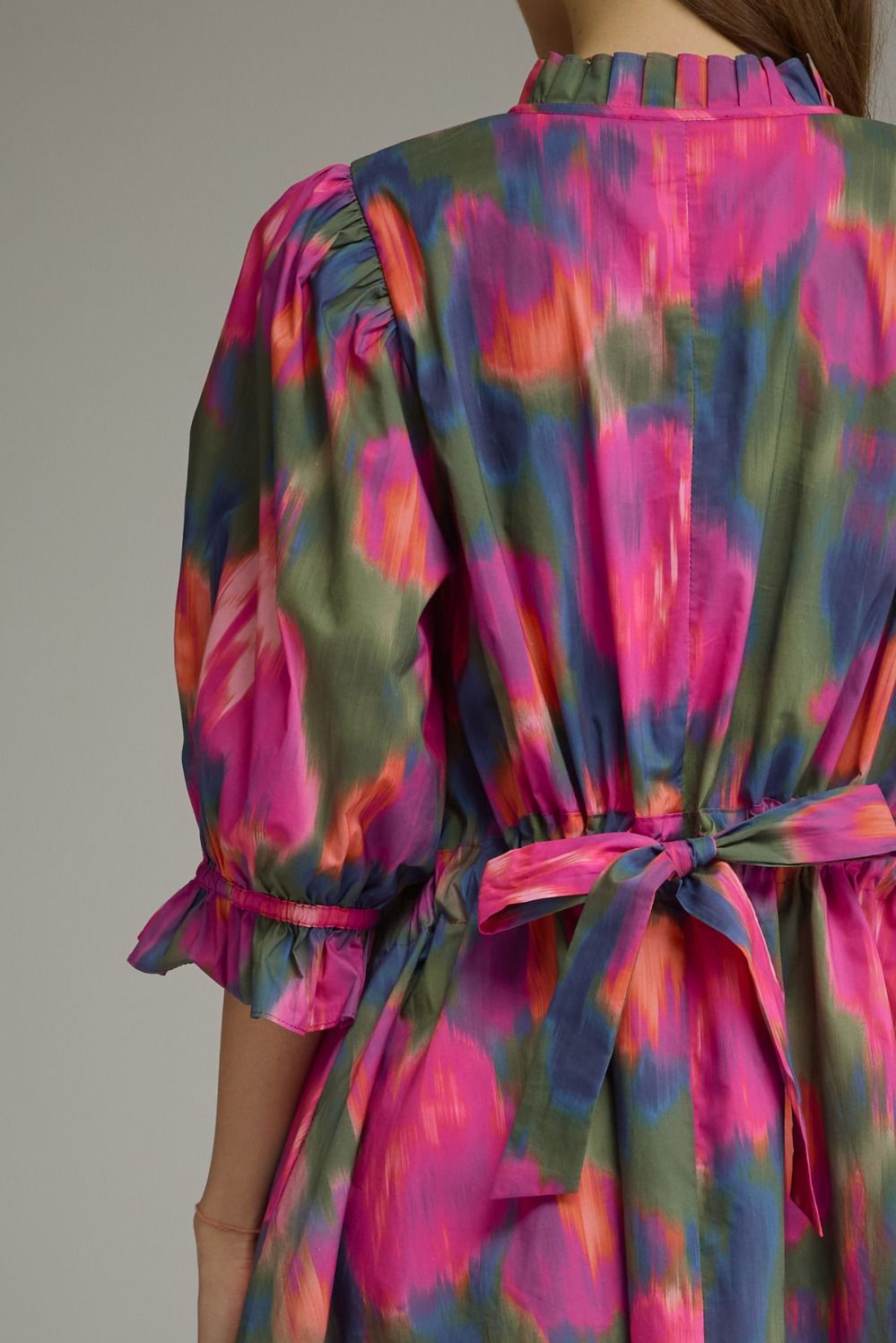 Close-up back view of a woman wearing a colorful dress with a pink, green, and blue abstract print, gathered waist tie, and puff sleeves.