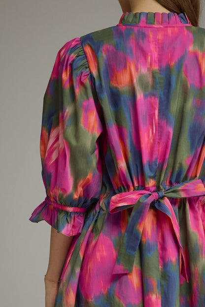 Close-up back view of a woman wearing a colorful dress with a pink, green, and blue abstract print, gathered waist tie, and puff sleeves.