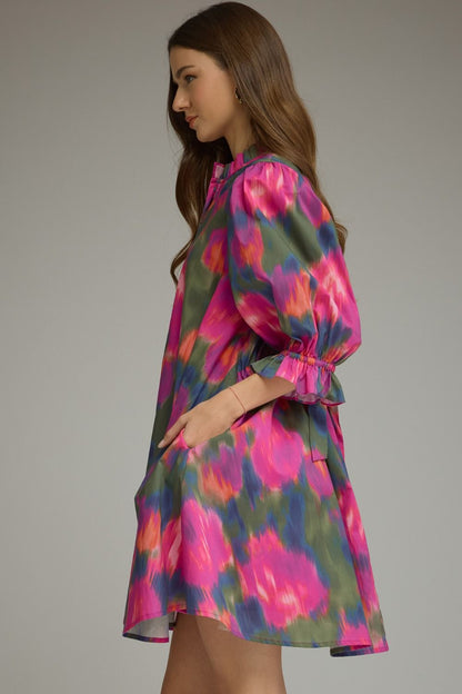 A woman wearing a short puff-sleeve dress with a vibrant pink, purple, and green watercolor floral pattern, shown from a side profile against a neutral gray studio background.