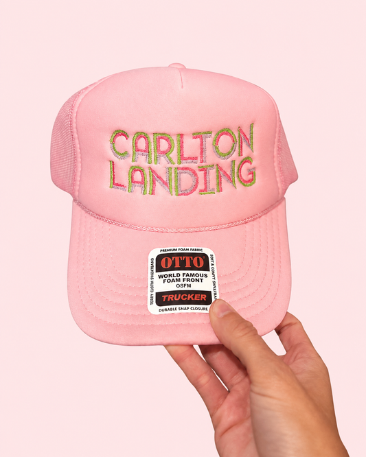A pink trucker-style cap with 'CARLTON LANDING' embroidered across the front in a multicolor gradient, held by a hand against a solid pale pink background. The cap features a structured foam front panel and mesh back, with a vintage-style patch on the brim.