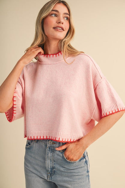 A woman wearing a light pink knit mock-neck sweater with orange scalloped trim on the collar and sleeve cuffs, paired with light-wash blue jeans, against a solid beige background.