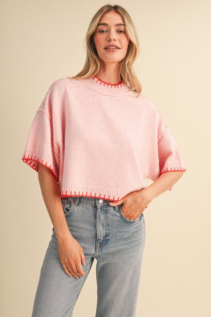 A woman modeling a light pink knit top with contrasting orange stitched trim along the neckline, sleeve hems, and bottom edge, paired with light-wash blue jeans against a solid beige background.