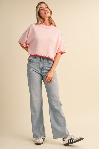 A woman modeling a light pink short-sleeve knit top with orange fringed trim, paired with light-wash wide-leg jeans and white sneakers, against a solid beige studio background.