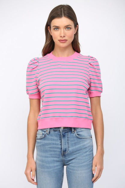 A woman wearing a pink short-sleeve knit top with thin horizontal blue stripes and puffed shoulders, paired with light-wash blue jeans, against a plain light gray studio background.