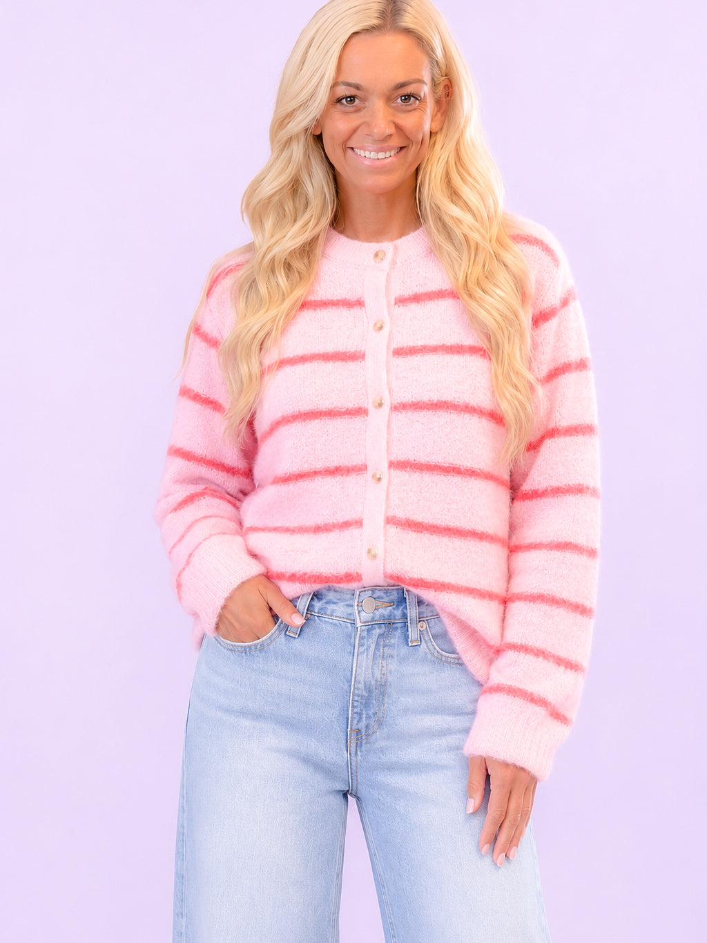 A woman with long blonde hair wearing a pink fuzzy cardigan with horizontal coral-pink stripes and light blue jeans, standing against a solid lavender background.
