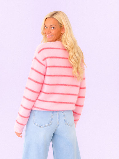 A blonde woman wearing a fuzzy pink and coral striped sweater and light blue jeans, photographed from the back against a solid light lavender background.