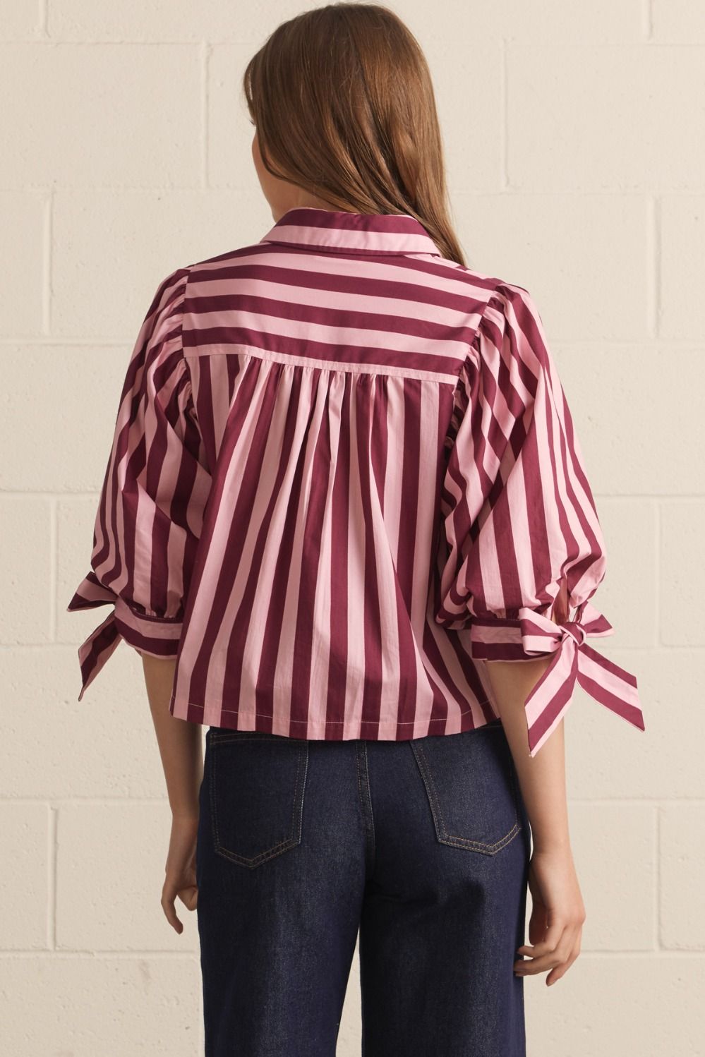 Person wearing a striped shirt with tied sleeves against a white brick wall.