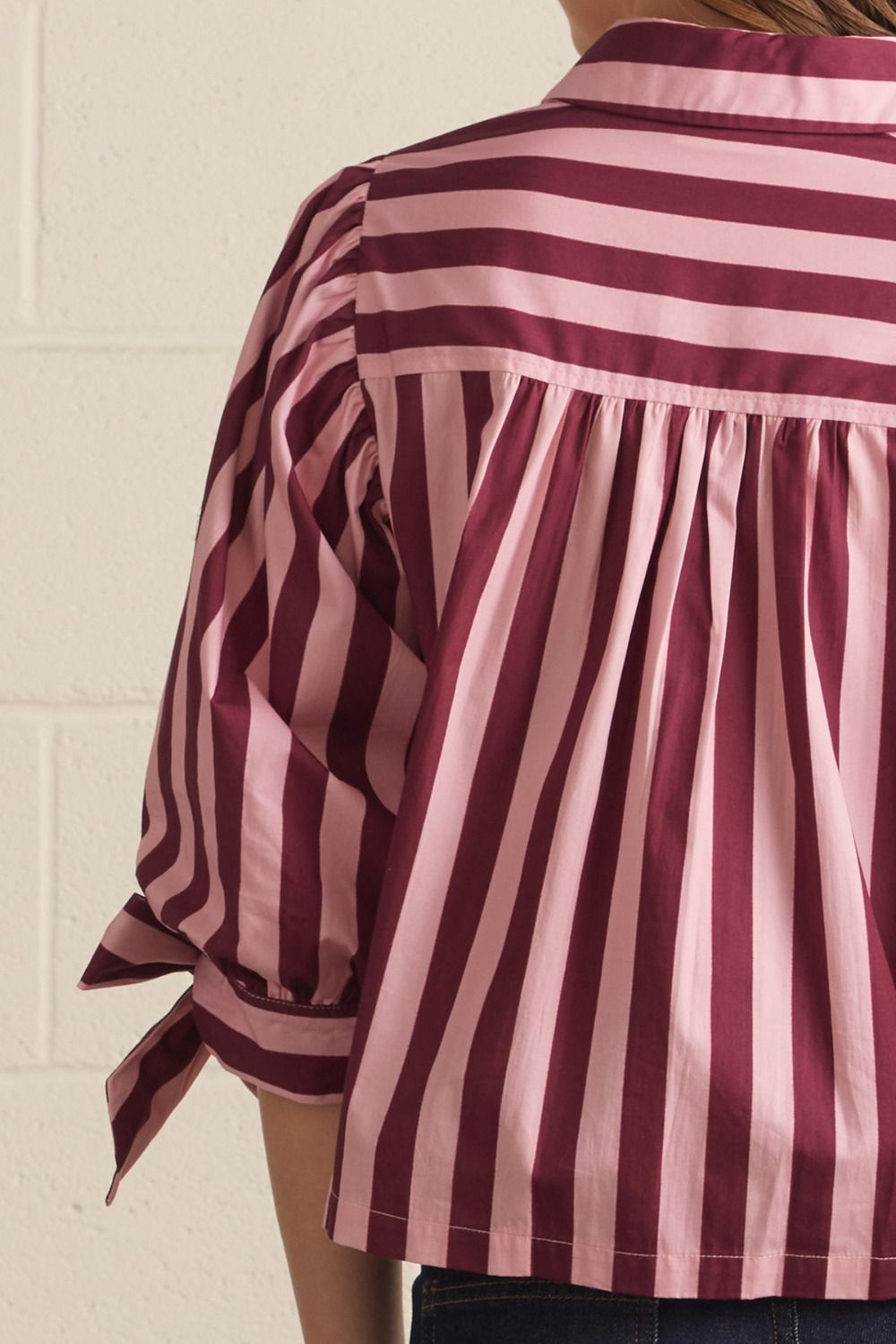 A close-up of a person wearing a pink and burgundy striped blouse, showing the back of the shirt with pleated detailing and rolled sleeves tied at the cuffs, set against a light brick wall.