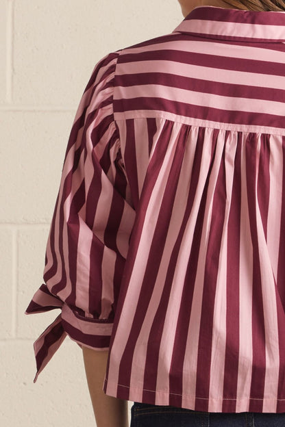 A close-up of a person wearing a pink and burgundy striped blouse, showing the back of the shirt with pleated detailing and rolled sleeves tied at the cuffs, set against a light brick wall.