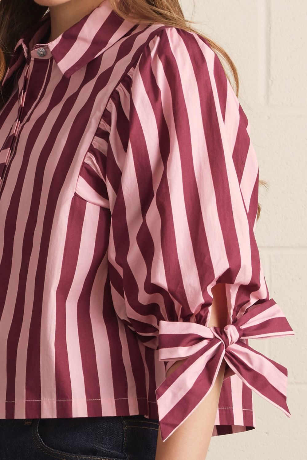 A close-up of a woman wearing a pink and burgundy striped blouse with puff sleeves and tied cuffs, paired with dark jeans against a light wall.