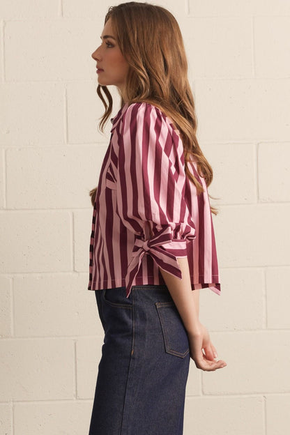 Woman wearing a pink and white striped shirt with a knot detail, standing against a beige tiled wall.