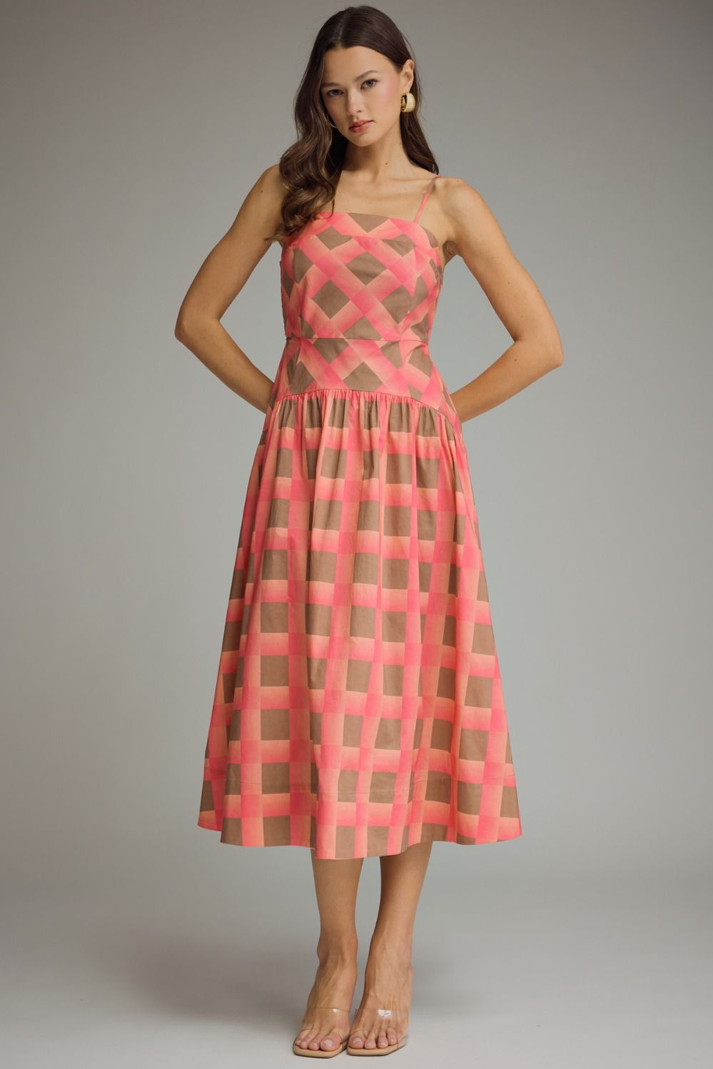 A woman modeling a sleeveless midi dress with a pink and green geometric pattern, standing against a neutral gray studio background.
