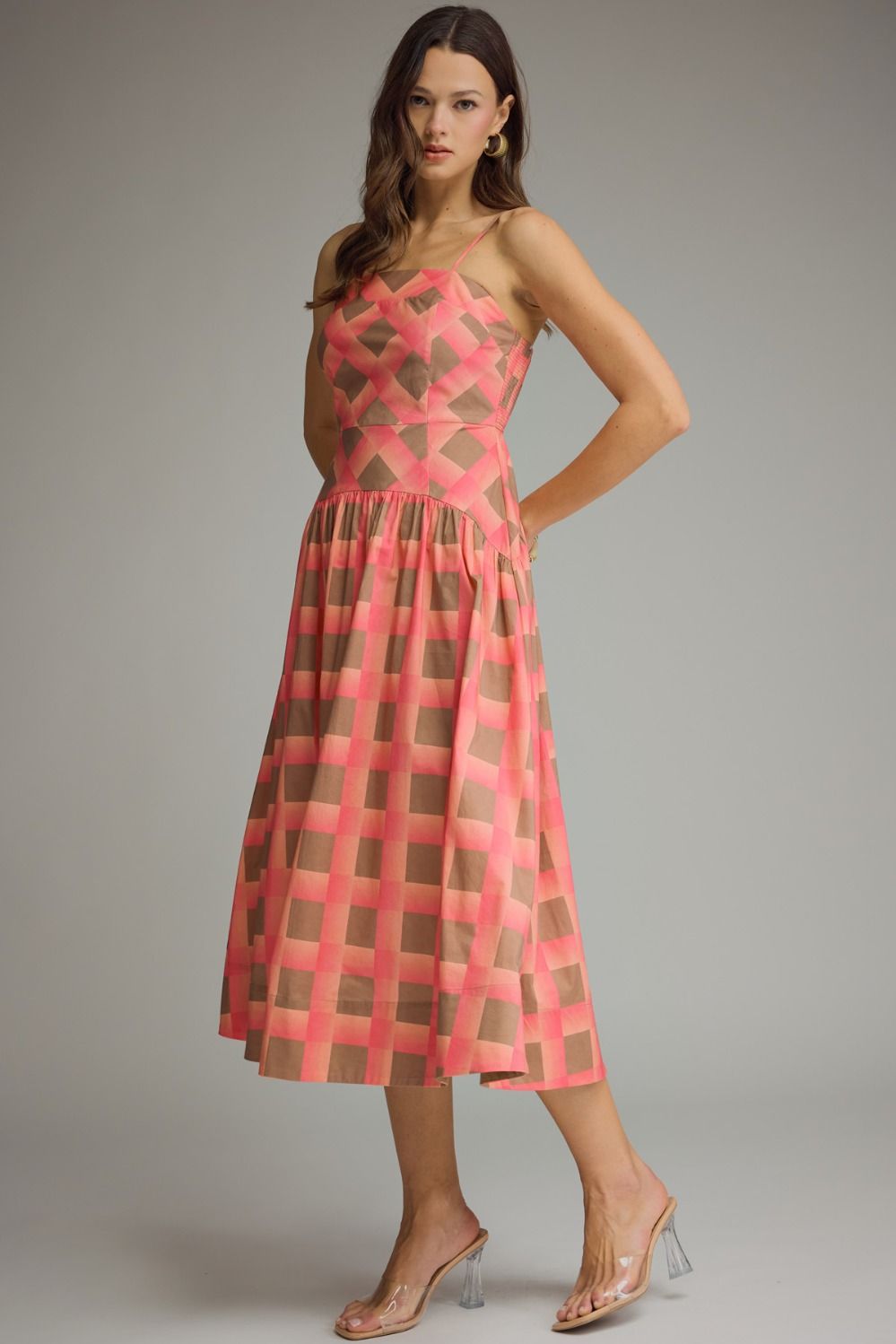 A woman models a sleeveless midi dress with a geometric pink and brown grid pattern against a plain gray studio background.