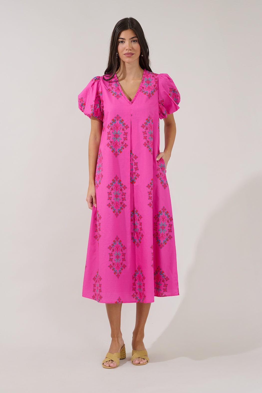 A woman modeling a bright pink midi dress with puff sleeves, a V-neckline, and a blue and green geometric pattern, set against a plain light gray studio background.