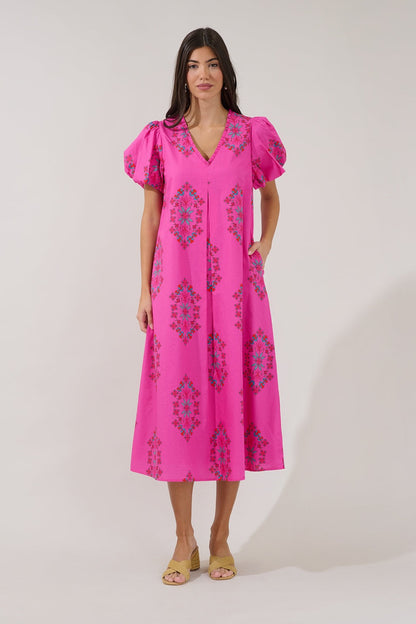 A woman modeling a bright pink midi dress with puff sleeves, a V-neckline, and a blue and green geometric pattern, set against a plain light gray studio background.