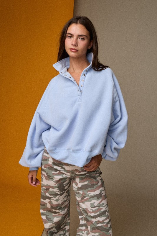 Woman wearing a light blue fleece pullover and camouflage pants against a two-tone wall.