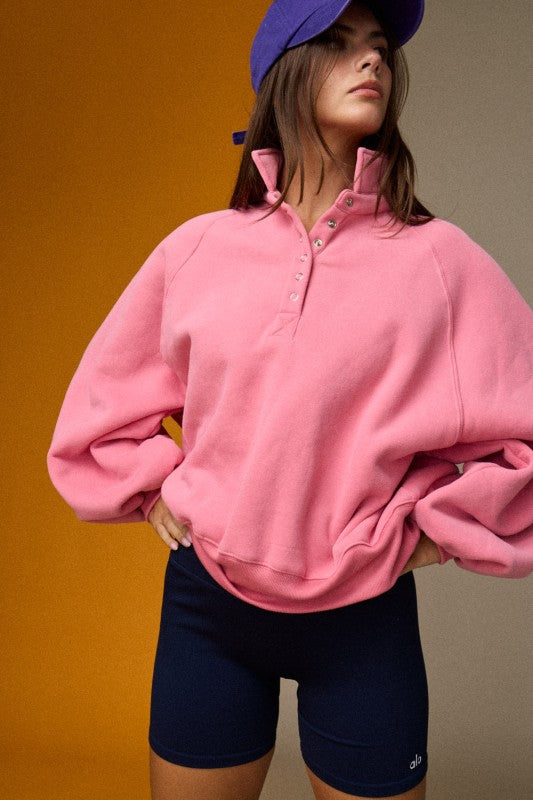 A woman wearing a pink quarter-zip sweatshirt with balloon sleeves, paired with navy blue bike shorts and a purple baseball cap, posing against a split orange and beige studio background.