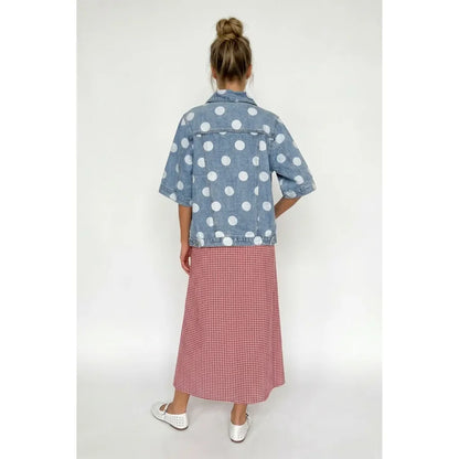 A woman shown from the back wearing a light blue denim shirt jacket with white polka dots and a pink and white gingham midi skirt, paired with white shoes, against a plain white studio background.