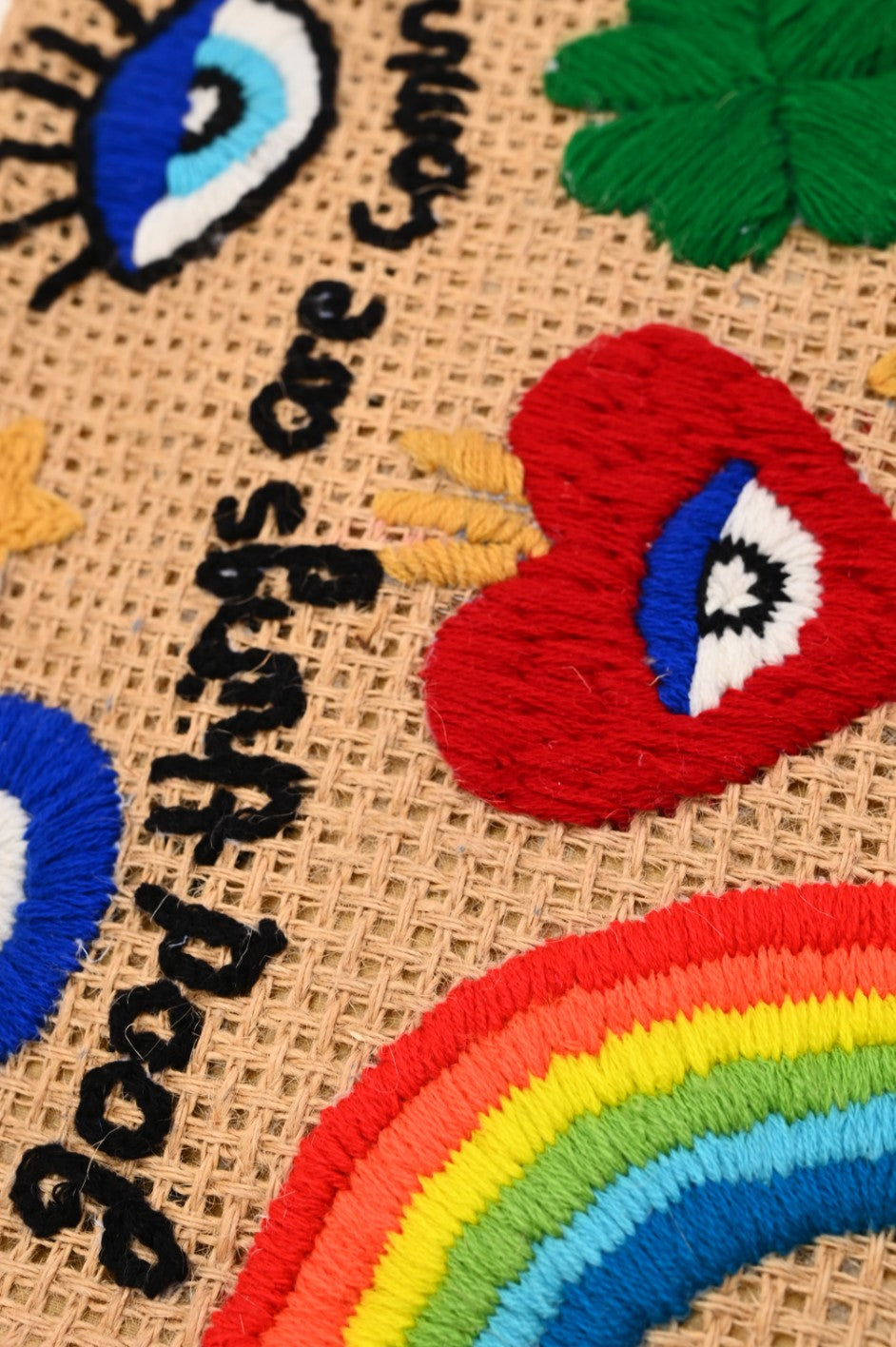 Natural woven tote bag with bamboo-style handles and colorful embroidered designs including a rainbow, heart, clover, eyes, and the phrase “good things are coming.”

