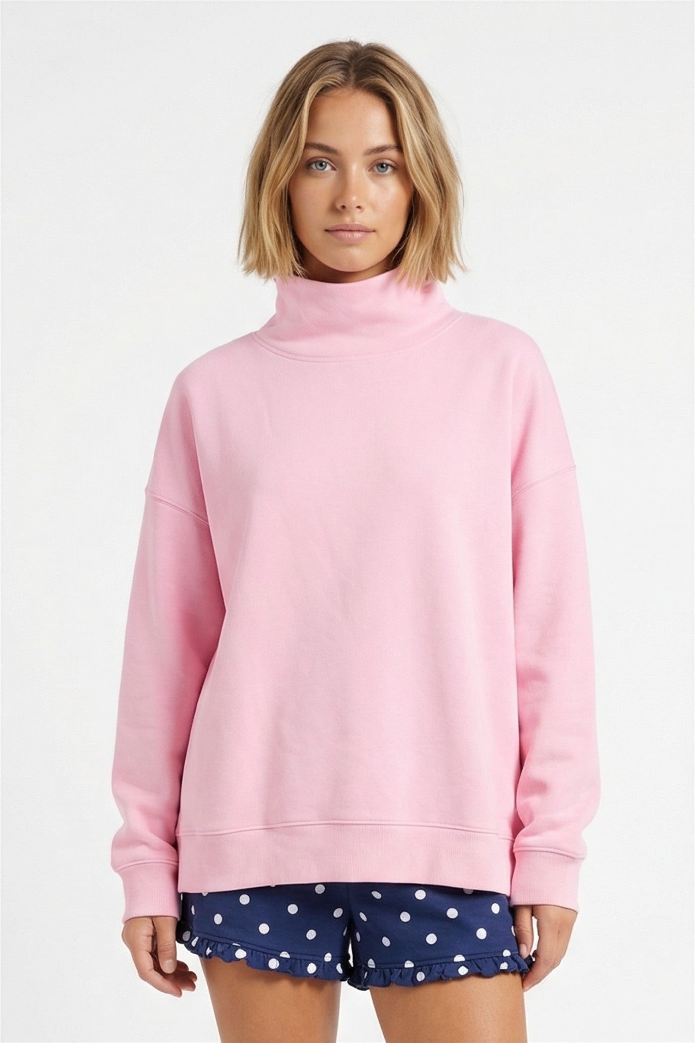 A woman wearing a light pink mock neck sweatshirt and navy blue polka dot shorts, standing against a plain white studio background.