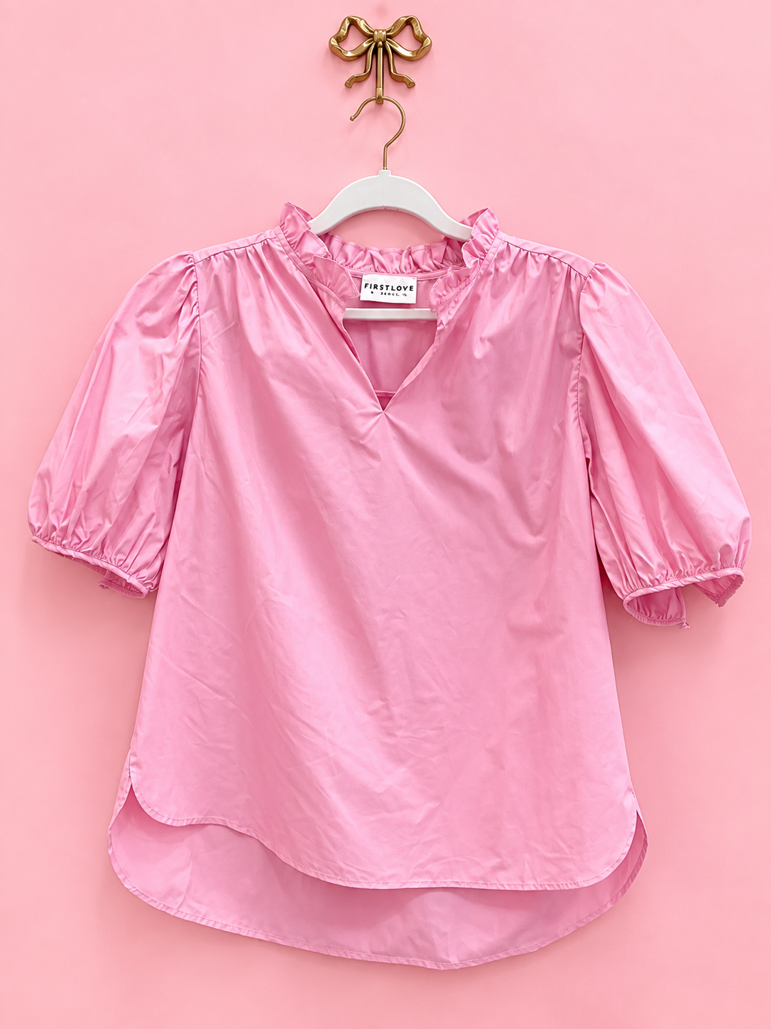 A pink short-sleeve blouse with a ruffled high neck and puff sleeves, hanging on a white hanger against a solid pink background.